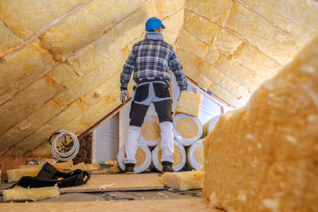 A worker wearing safety gear stands in an attic surrounded by rolls of insulation material. Sunlight filters through the roof, illuminating the unfinished space. The worker carefully inspects the area, preparing to enhance the home's energy efficiency by installing insulation in the ceiling.
